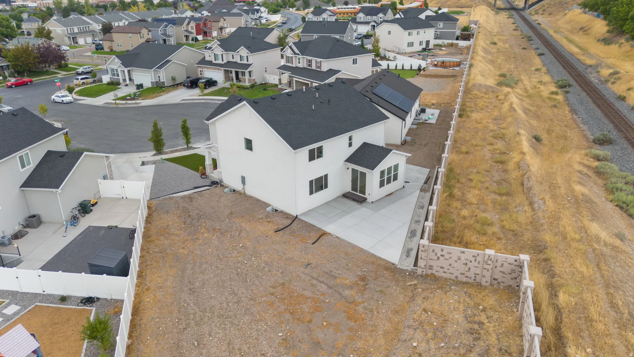 Expansive rear yard with a newly poured concrete patio, a retaining wall, and a newly constructed home with a dark shingle roof and solar panels