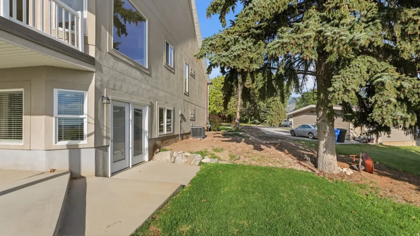Stucco exterior with multiple windows, concrete patio, glass panel double doors, and a grass lawn with mature trees