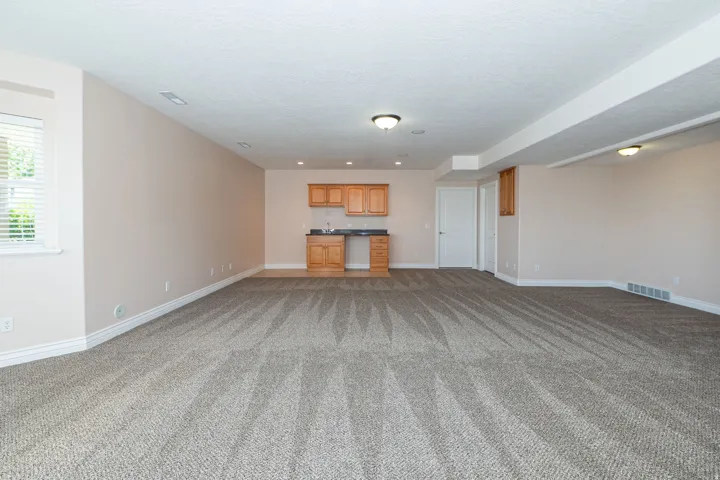Spacious room featuring neutral carpeting, a built-in wet bar with wood cabinetry and a dark countertop, recessed lighting, and a window with blinds