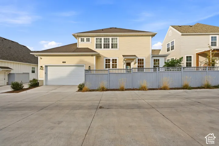 Cream-colored siding exterior featuring multiple windows with white trim, a two-car garage with a white door, a gray privacy fence with lattice detail, and a concrete driveway