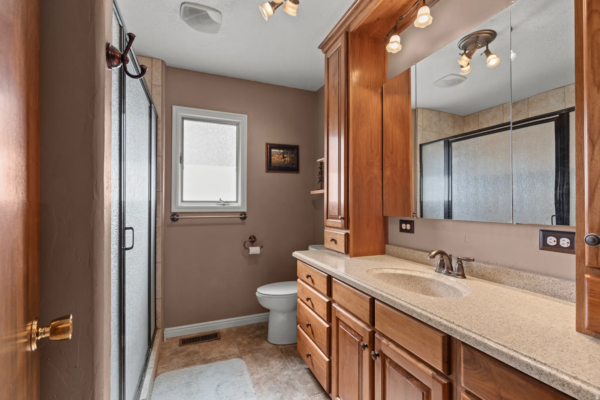 Bathroom featuring a glass-enclosed shower, a single vanity with an integrated sink and stone-finish countertop, wood cabinetry with ample storage, a window, and a toilet