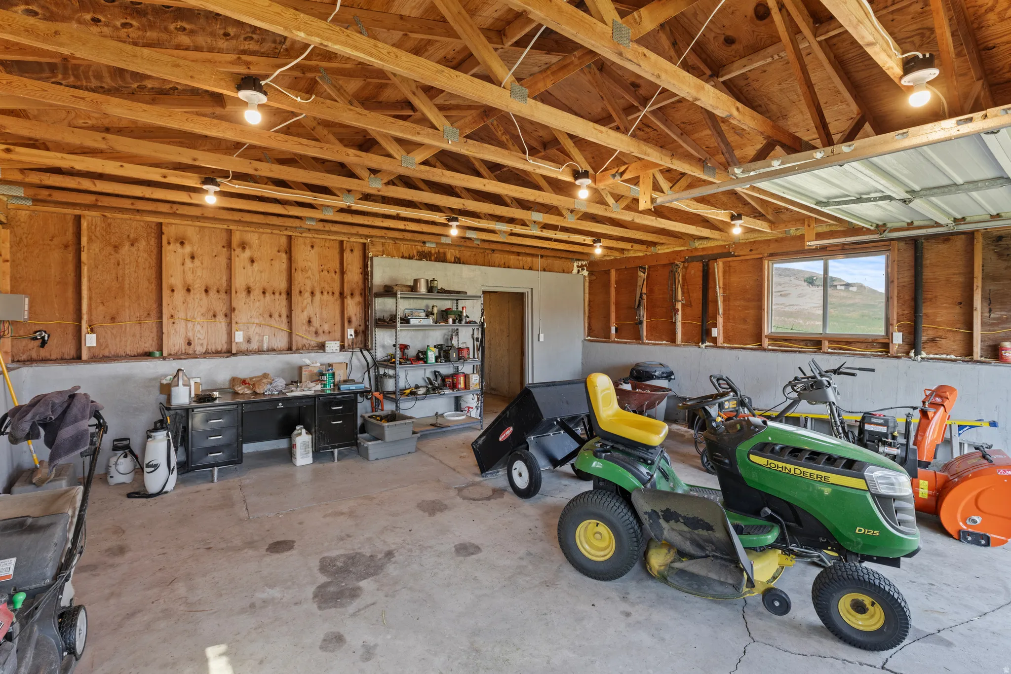 Spacious 1 car garage featuring exposed wood beam ceiling, concrete flooring, ample work surfaces, built-in shelving, and multiple overhead light fixtures