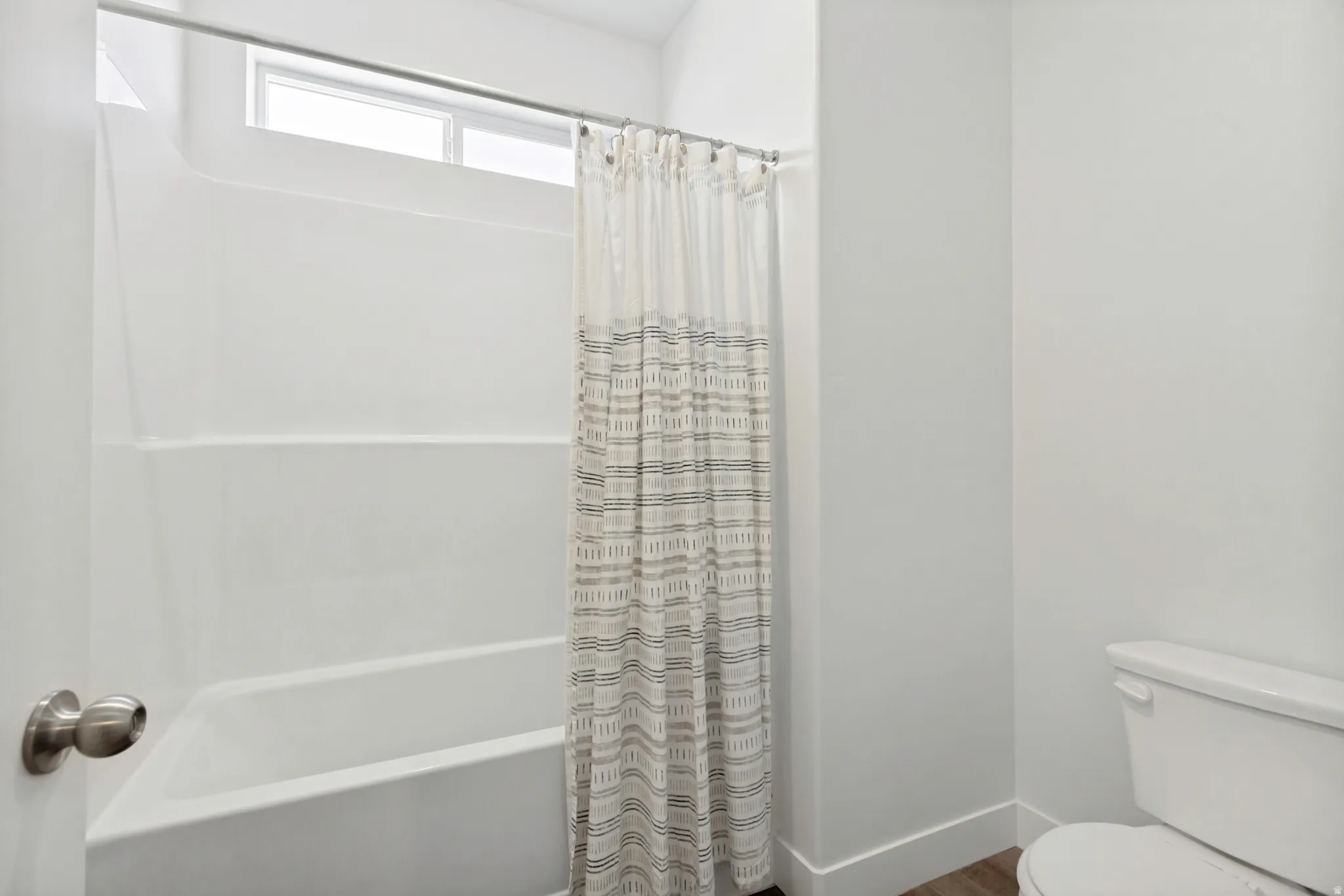 White bathtub and shower combination unit featuring a transom window, brushed nickel shower rod, white walls, and wood-finish flooring