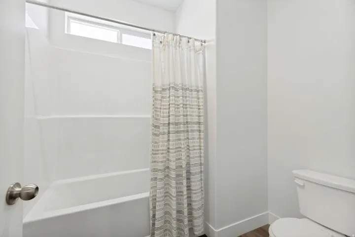 White bathtub and shower combination unit featuring a transom window, brushed nickel shower rod, white walls, and wood-finish flooring