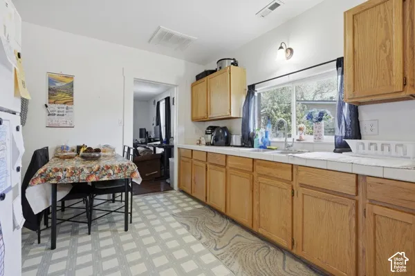 Wood-finish cabinetry, tile countertops, stainless steel sink, large window, and patterned flooring