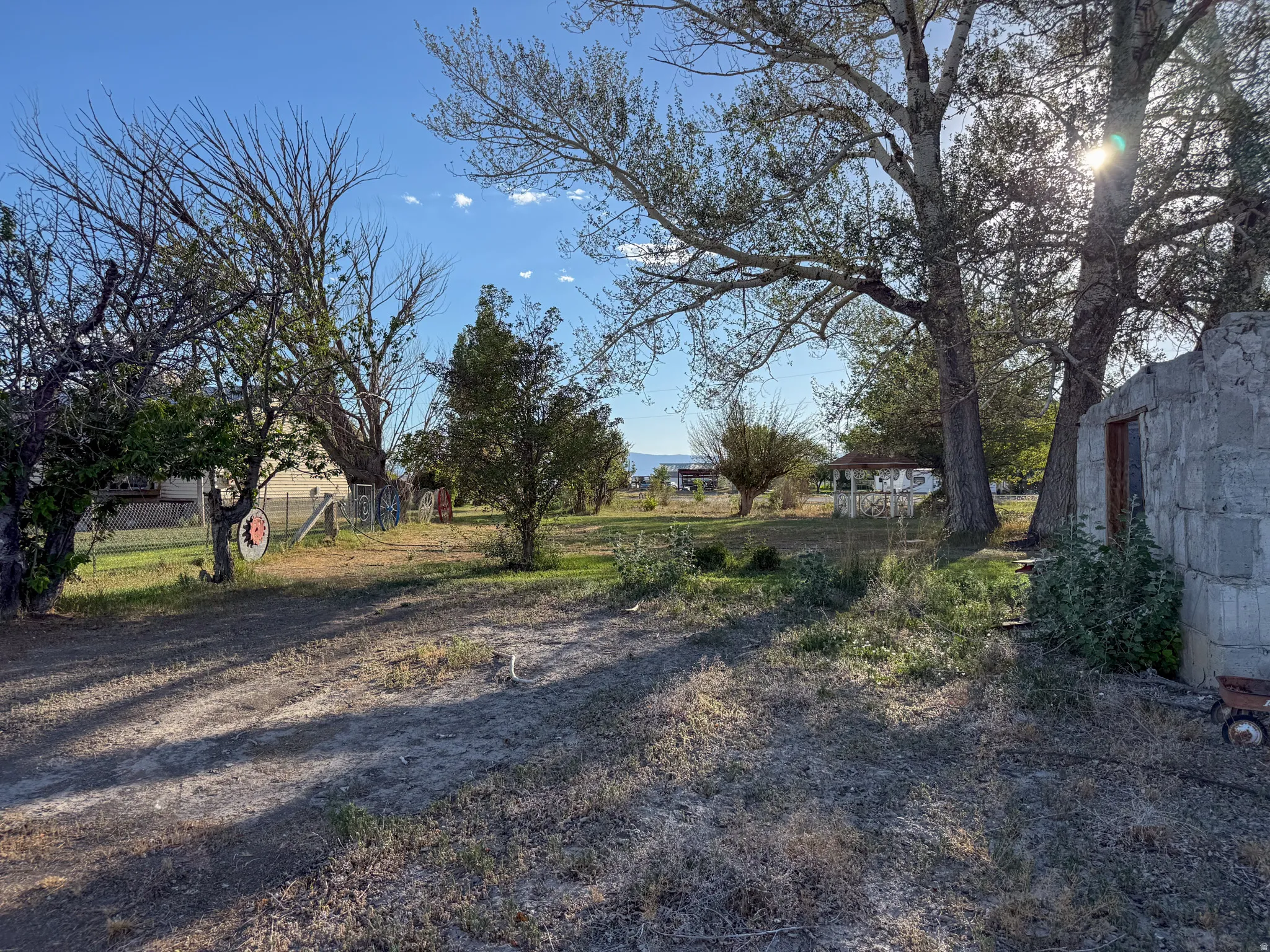 Expansive exterior space featuring mature trees providing natural shade, a clear blue sky, distant mountain range, a partially visible white gazebo, and a stone outbuilding