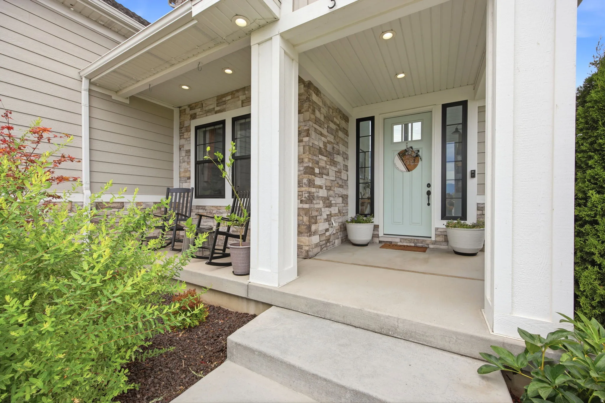 Inviting front porch featuring a light blue entry door with flanking sidelights, stone veneer accents, recessed lighting, and a concrete entry pad with steps