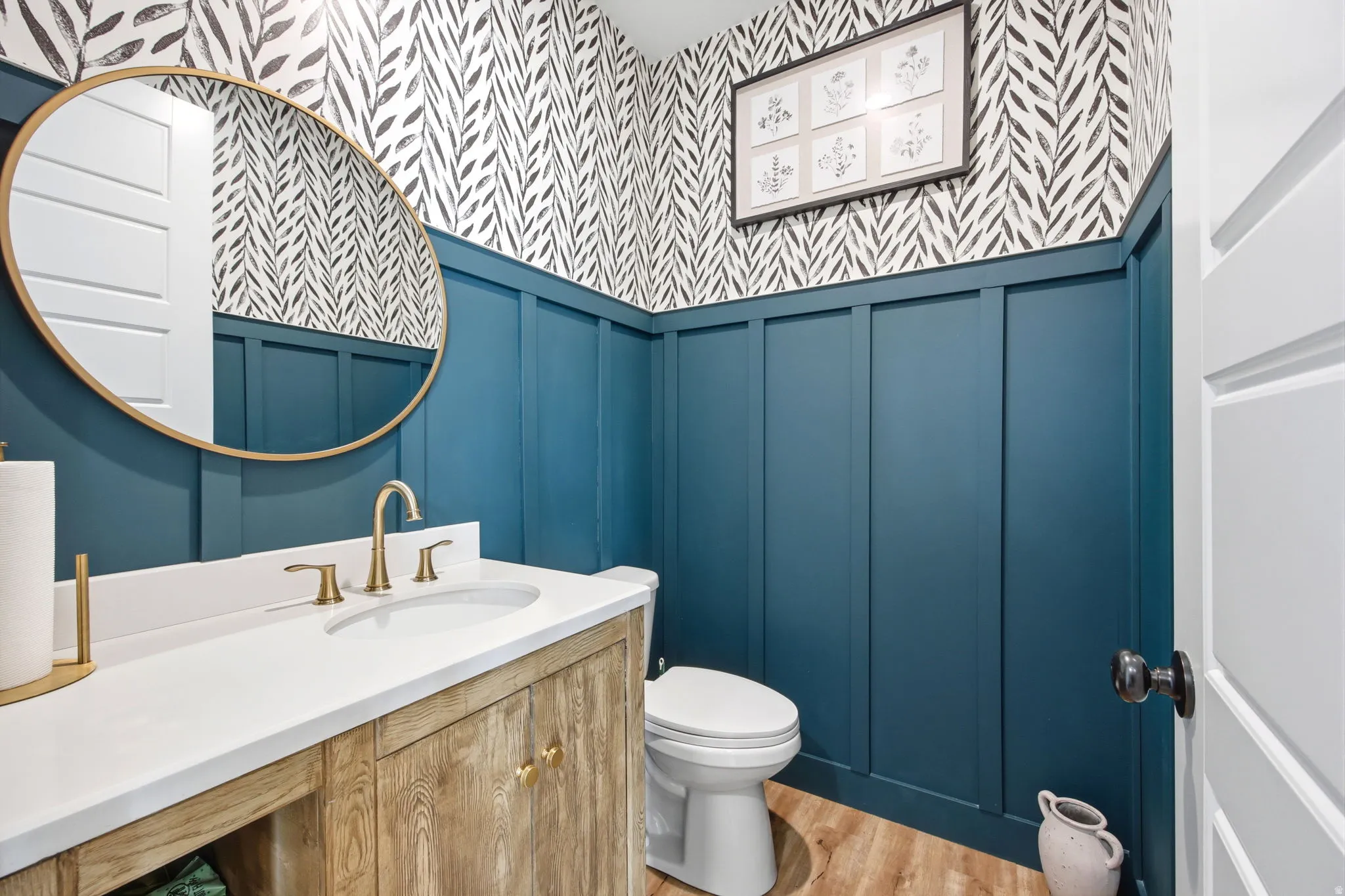Powder room featuring board and batten wainscoting, decorative wallpaper, a wood-finish vanity with a white countertop, and gold-finish fixtures