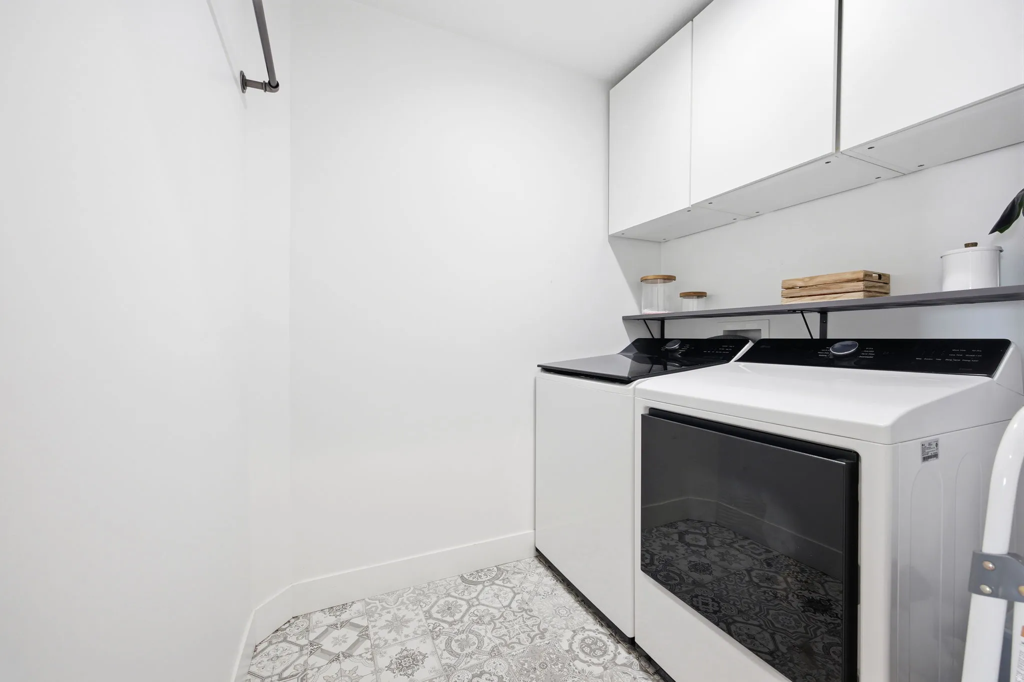 Dedicated laundry area featuring patterned tile flooring, overhead white cabinetry, and a dark floating shelf
