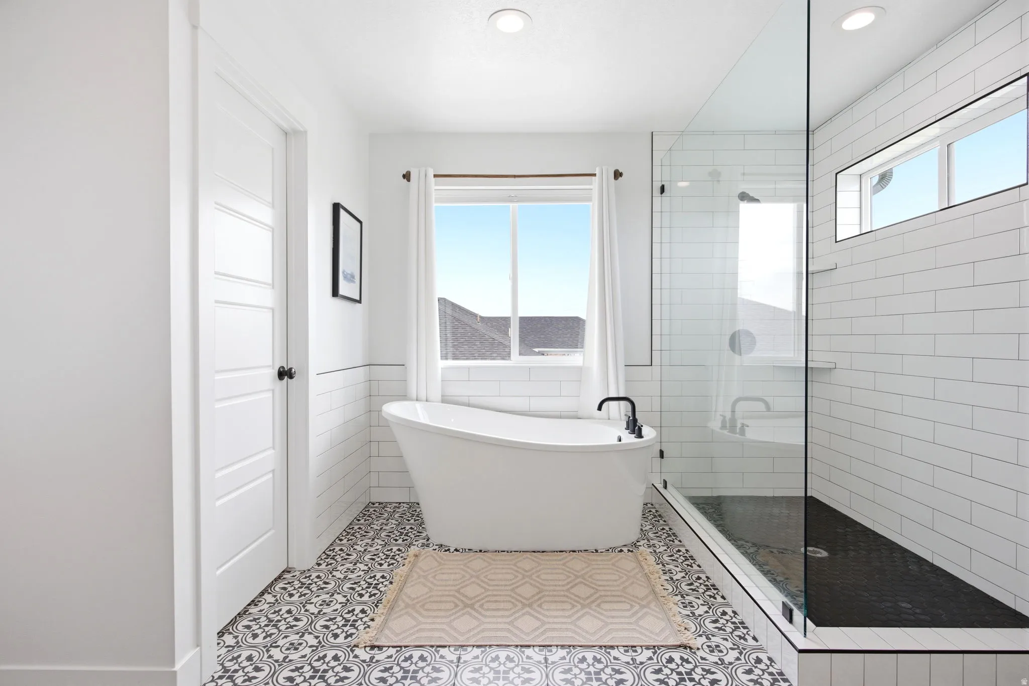 Bathroom featuring a freestanding soaking tub with a matte black faucet, a glass-enclosed shower with subway tile surround, patterned floor tiles, and a window with white curtains