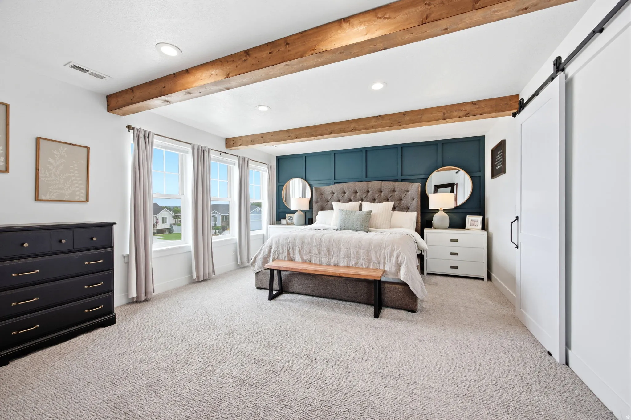 Spacious bedroom featuring exposed wood beams, recessed lighting, a decorative accent wall, three large windows, and a sliding barn door