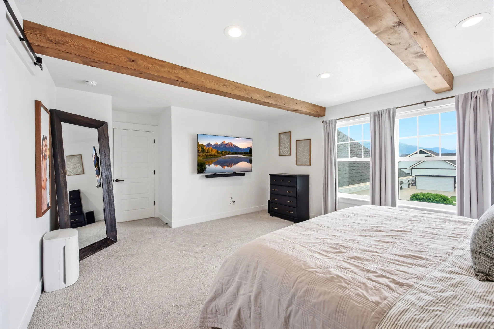 White-painted interior space featuring exposed wood ceiling beams, two windows with light-colored drapes, recessed lighting, and light-colored carpet flooring