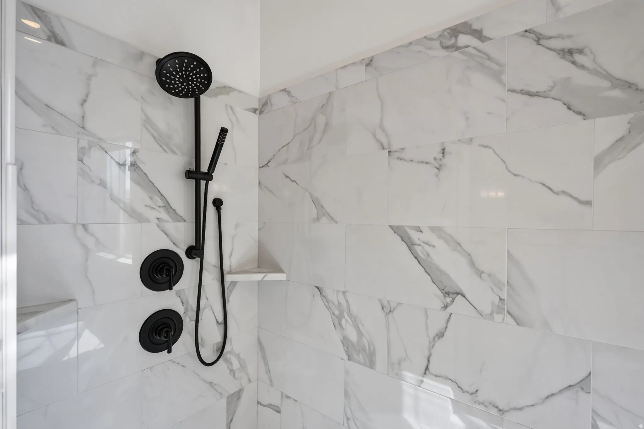 Shower with large format white and gray veined tile, matte black shower fixtures, hand-held shower, and an integrated corner shelf