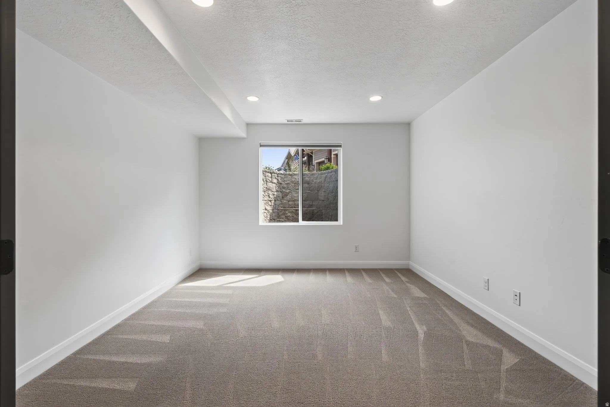 Spacious room featuring neutral carpeting, white walls, recessed lighting, and a window with natural light