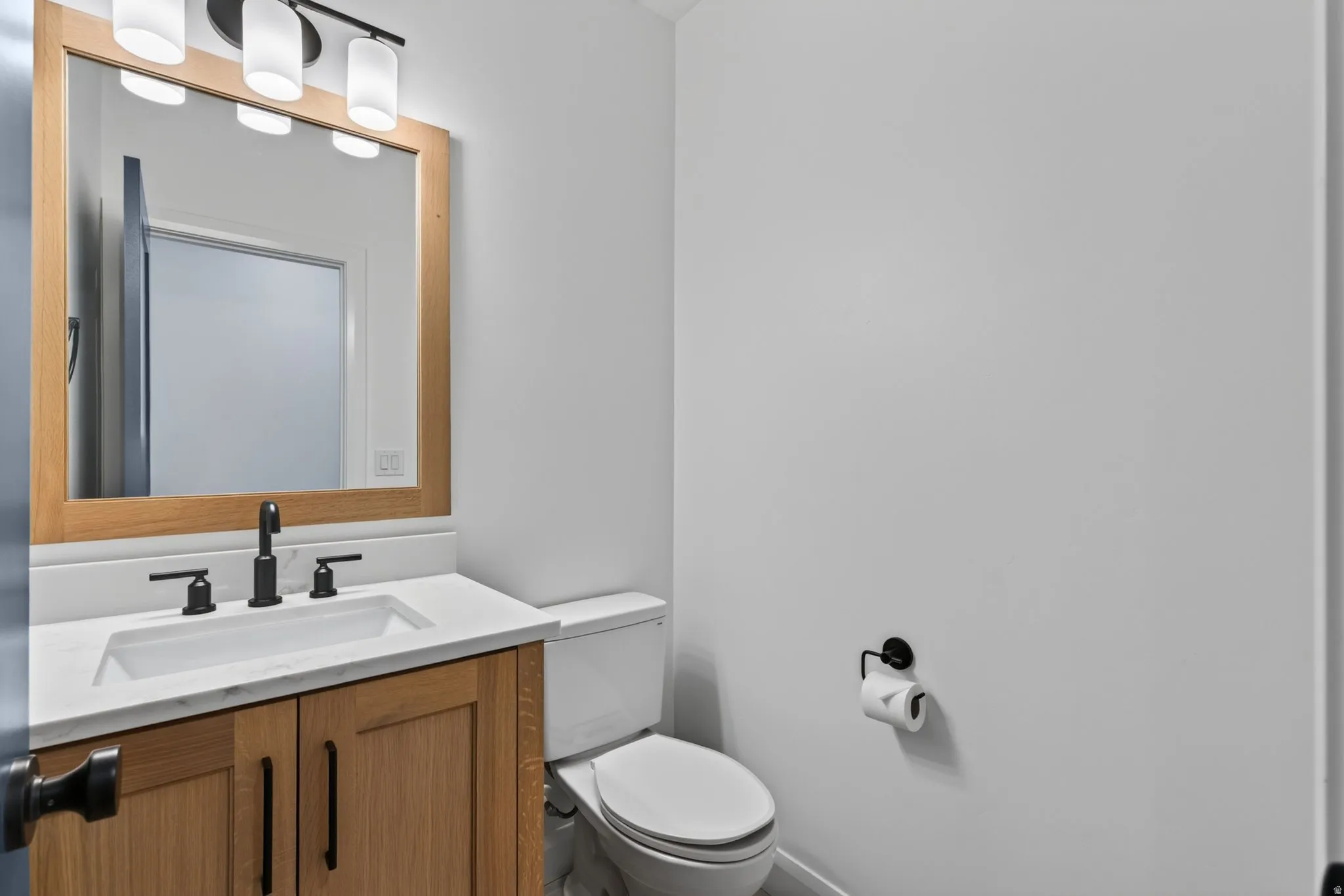 Contemporary vanity featuring a light wood-finish cabinet with black hardware, a white countertop with an integrated rectangular sink, and a black faucet
