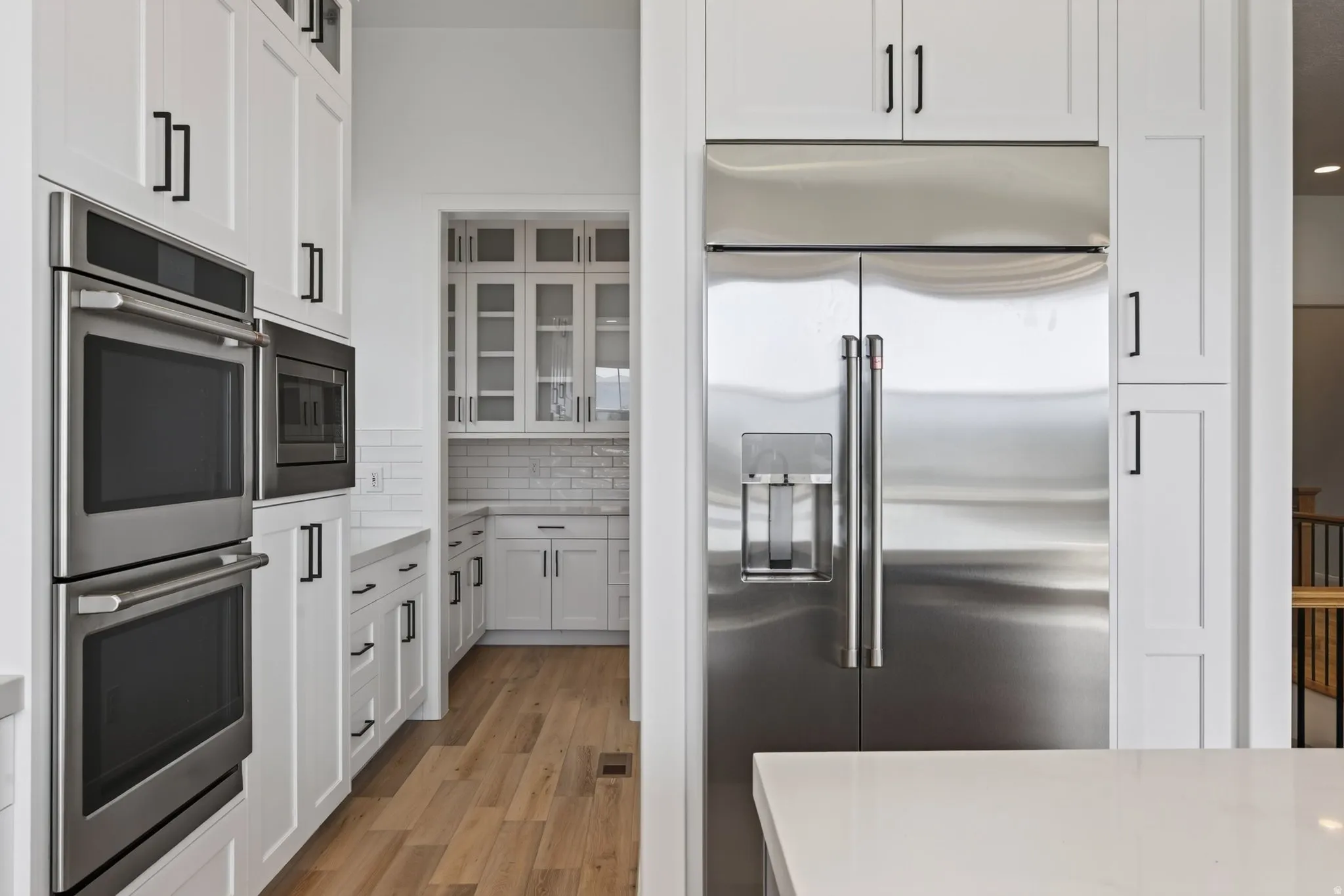 Kitchen featuring white cabinetry, stainless steel appliances, light countertops, wood-finish flooring, and a walk-in pantry with glass-front upper cabinets and subway tile backsplash