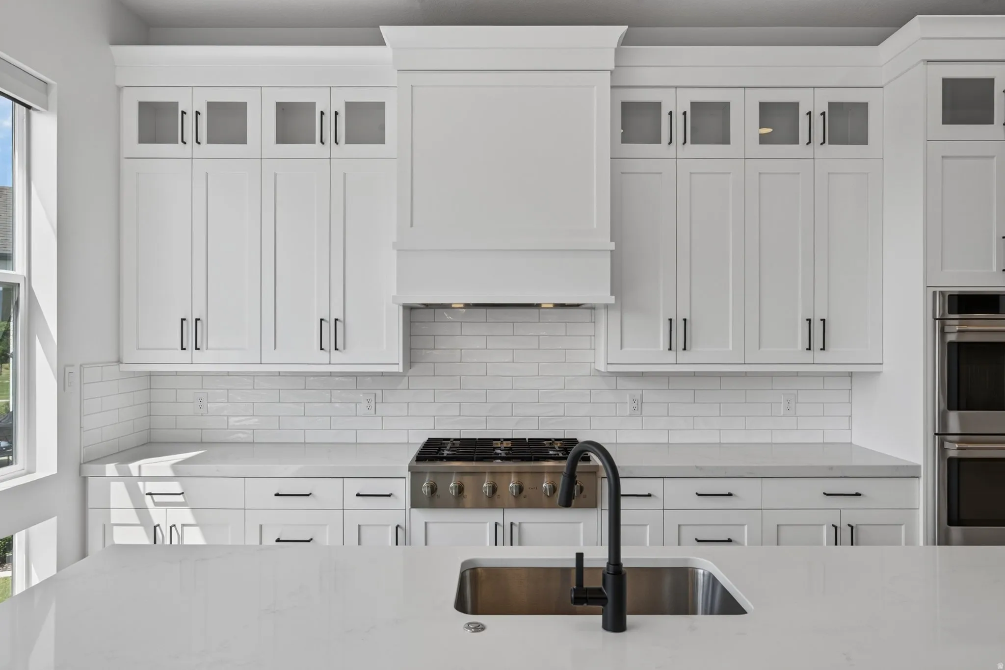 Modern kitchen featuring white shaker cabinetry with upper glass-front cabinets, a built-in range hood, subway tile backsplash, and light-toned countertops