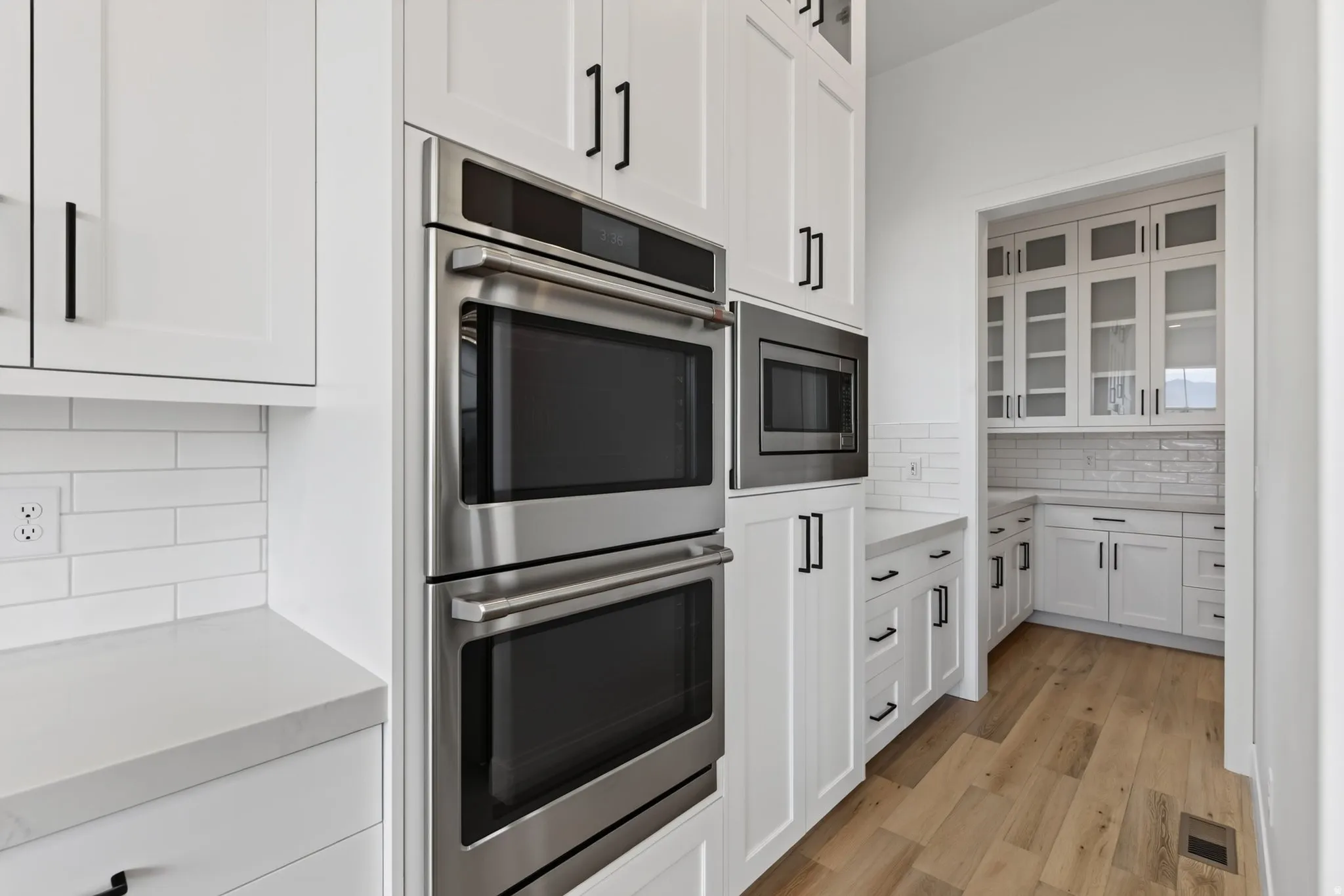 Modern kitchen featuring white cabinetry with black hardware, stainless steel double ovens and microwave, light-toned countertops, and subway tile backsplash