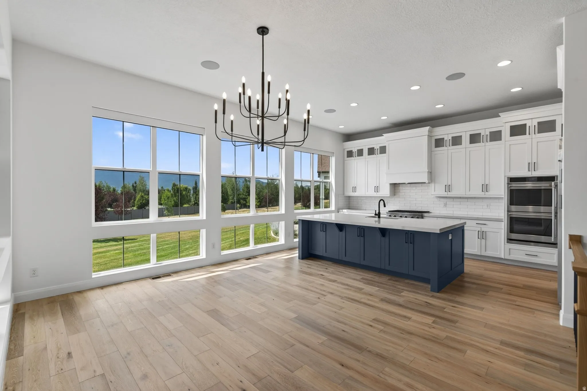 Open-concept kitchen and dining area featuring a large center island with a sink, white cabinetry with upper glass-front accents, and stainless steel double ovens