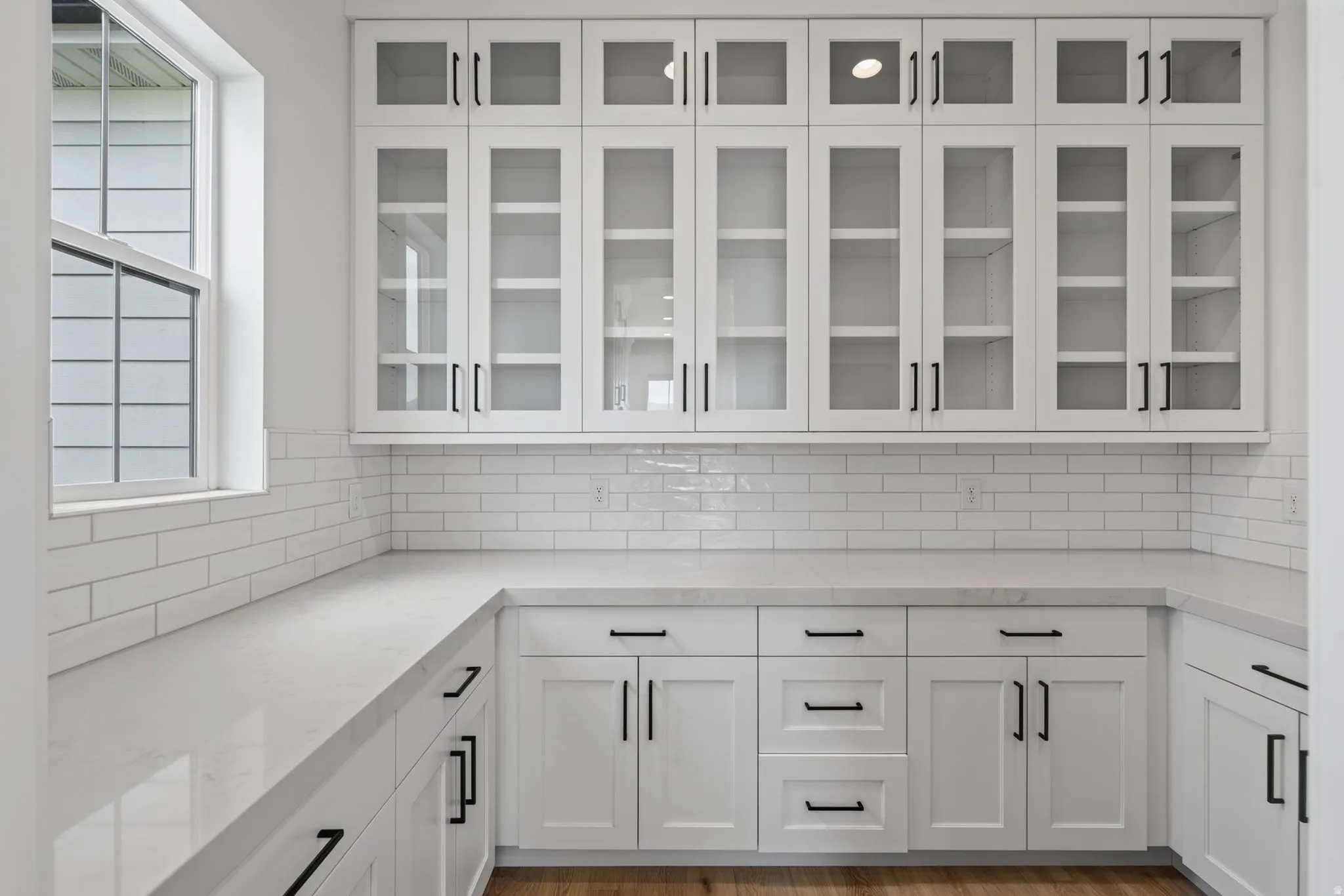 Modern kitchen featuring extensive white cabinetry with matte black hardware, glass-front upper cabinets, light-toned countertops, and a white subway tile backsplash