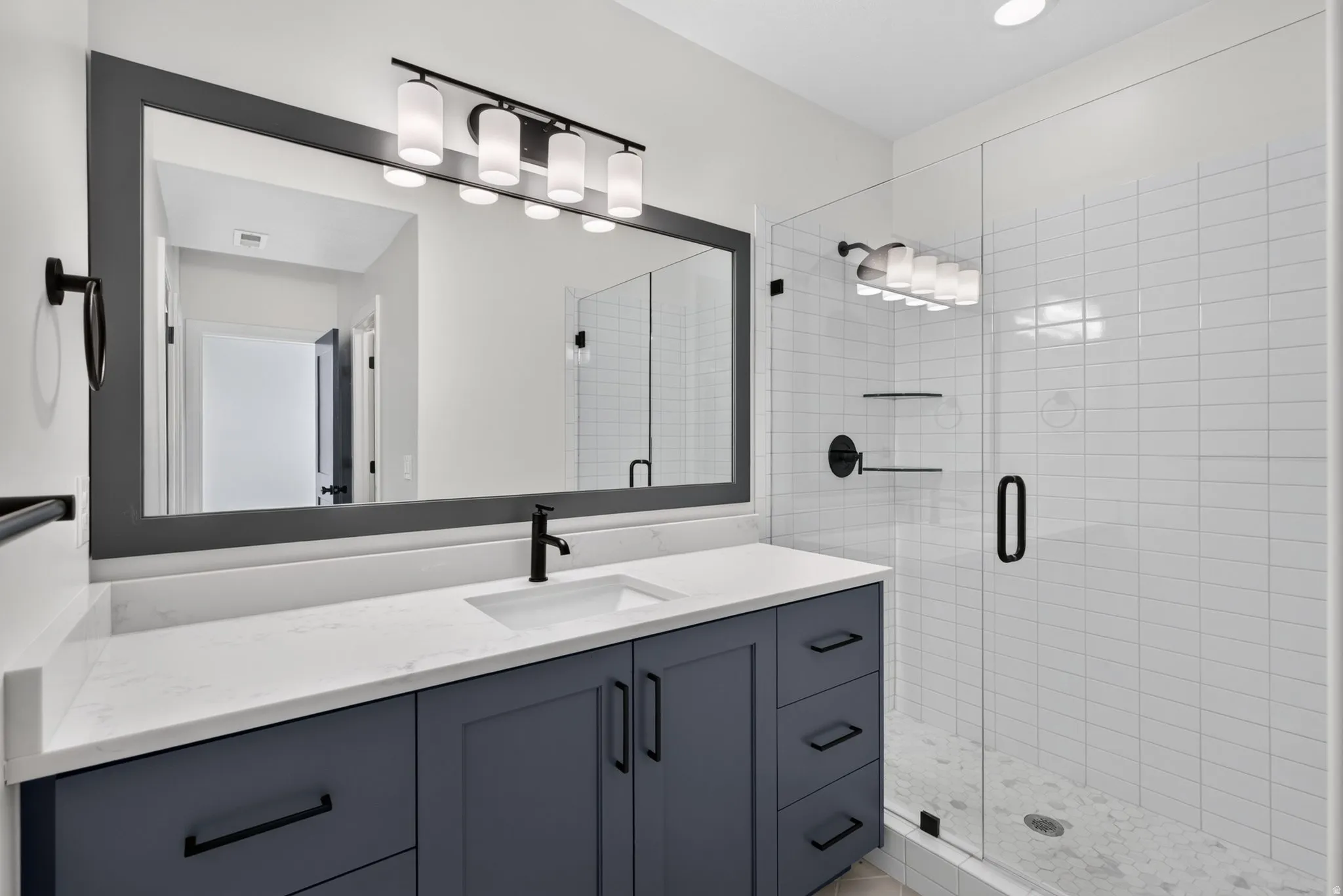 Bathroom featuring a dark blue vanity with a white countertop and integrated sink, a rectangular framed mirror, a five-light vanity fixture, and a glass-enclosed shower with white subway tile and a pebbled floor