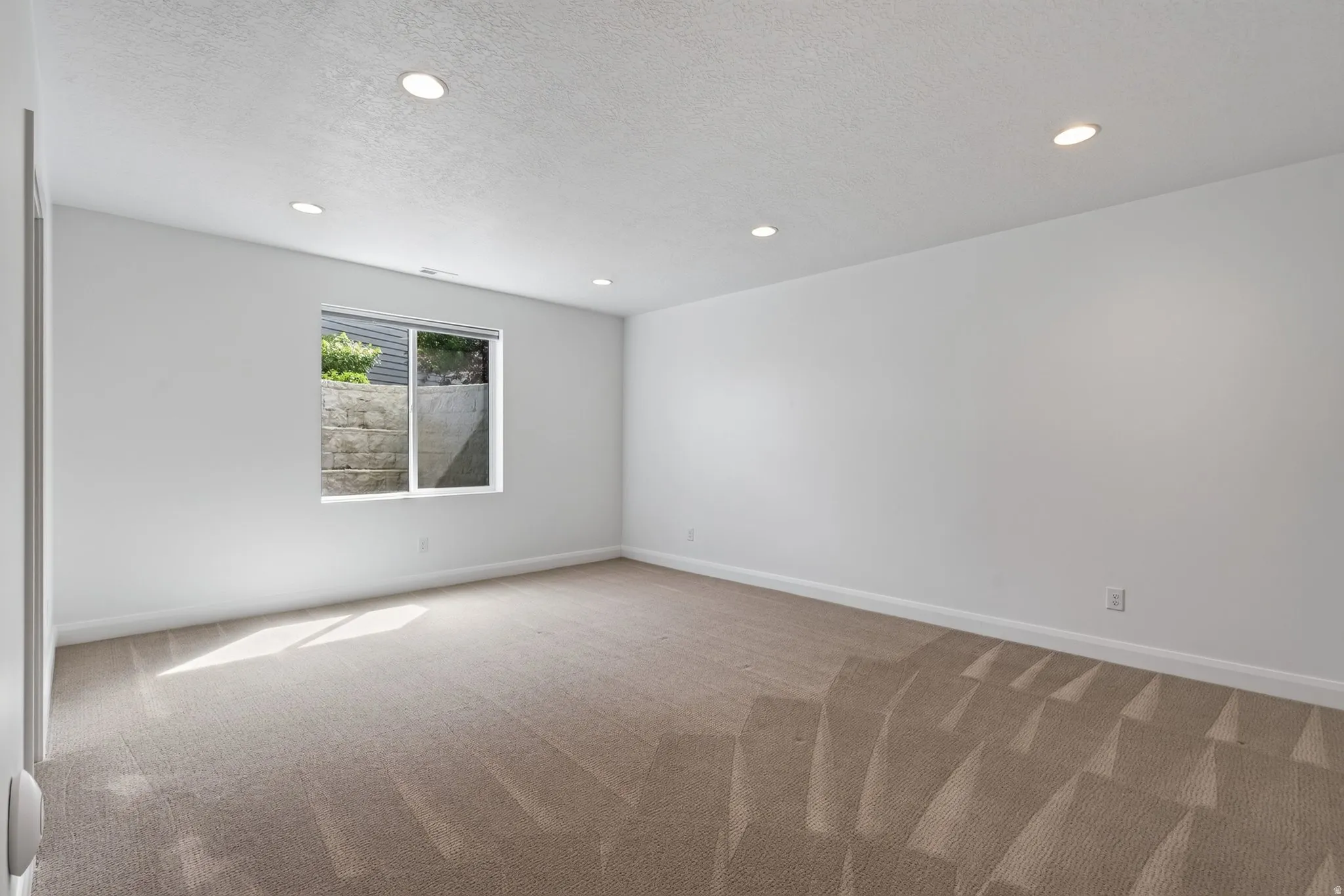 Spacious room featuring neutral carpet flooring, recessed lighting, white walls, and a window with a stone retaining wall outside