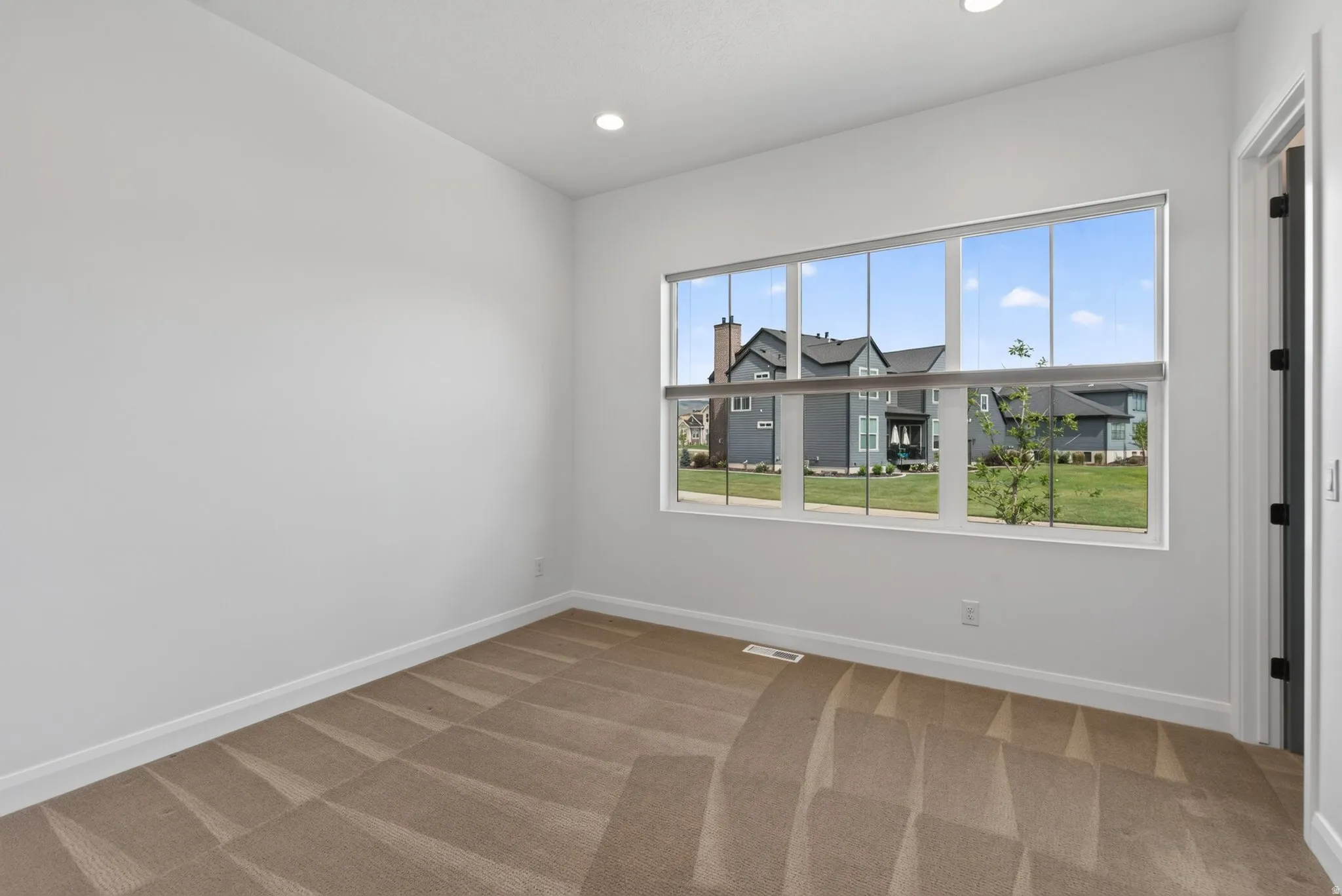 Recessed lighting and a multi-pane window illuminate this room, featuring light gray walls, white baseboards, and a patterned carpet