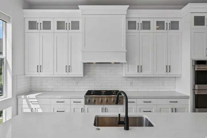 Modern kitchen featuring white shaker cabinetry with upper glass-front cabinets, a built-in range hood, subway tile backsplash, and light-toned countertops
