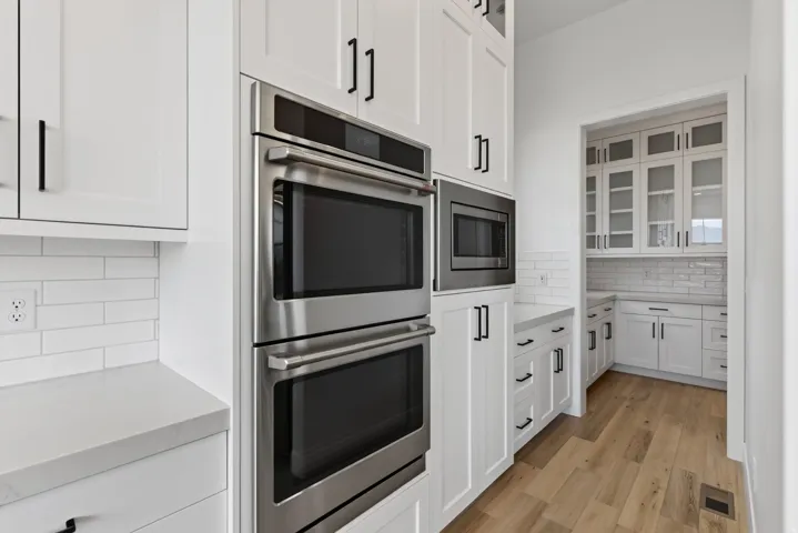 Modern kitchen featuring white cabinetry with black hardware, stainless steel double ovens and microwave, light-toned countertops, and subway tile backsplash