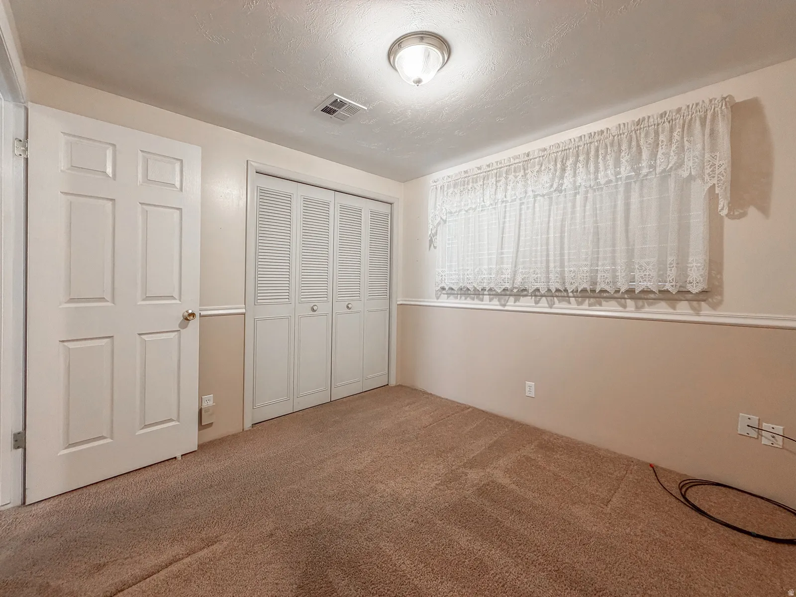 Carpeted room featuring a ceiling-mounted light fixture, louvered bi-fold closet doors, a four-panel interior door, and a horizontal window