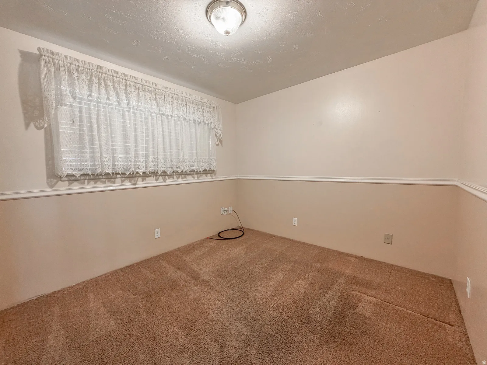 Carpeted room featuring a ceiling-mounted light fixture and a window with white lace curtains