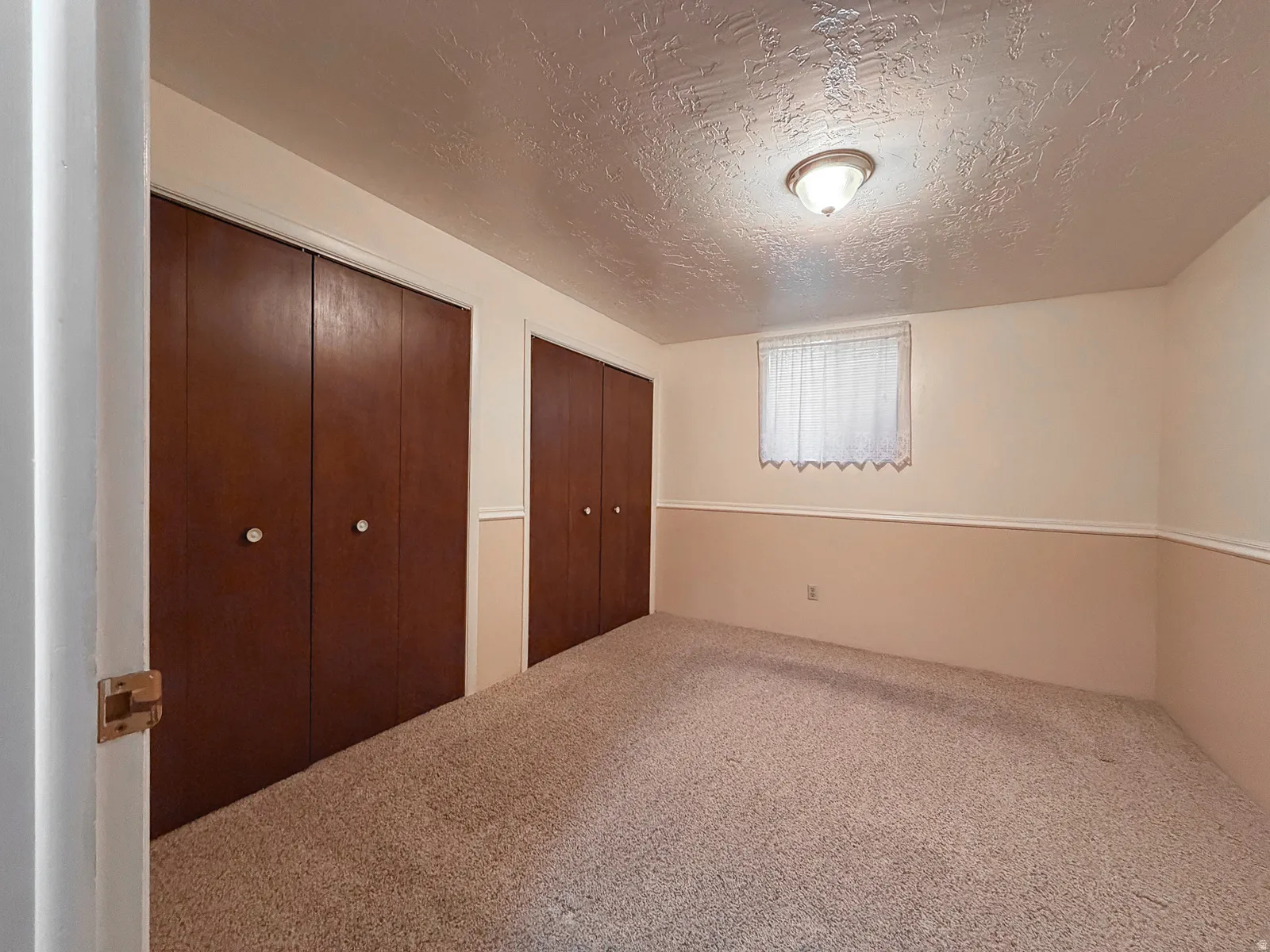 Carpeted room featuring two sets of bi-fold closet doors, a window with a sheer curtain, and a ceiling-mounted light fixture
