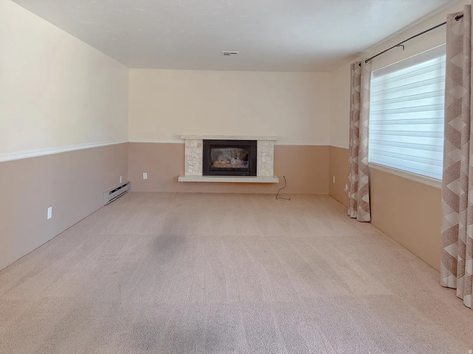 Living area featuring a fireplace with a light-toned stone surround, neutral carpet flooring, and a large window with blinds and patterned drapes