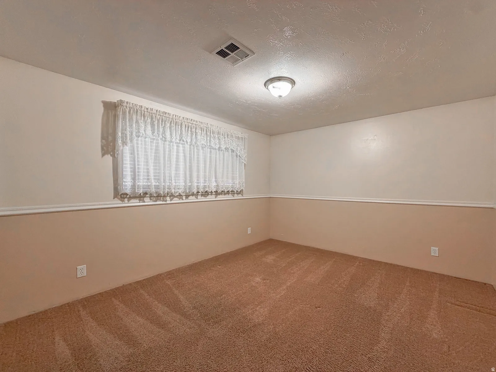Carpeted interior space featuring a window with white lace curtains, two-tone wall treatment, and an overhead flush-mount light fixture