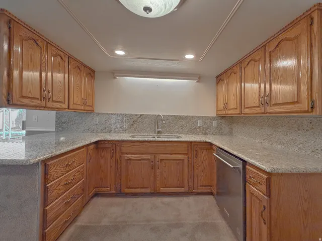 U-shaped kitchen featuring wood cabinetry, light-toned countertops with a matching backsplash, a stainless steel dishwasher, and a double-basin sink