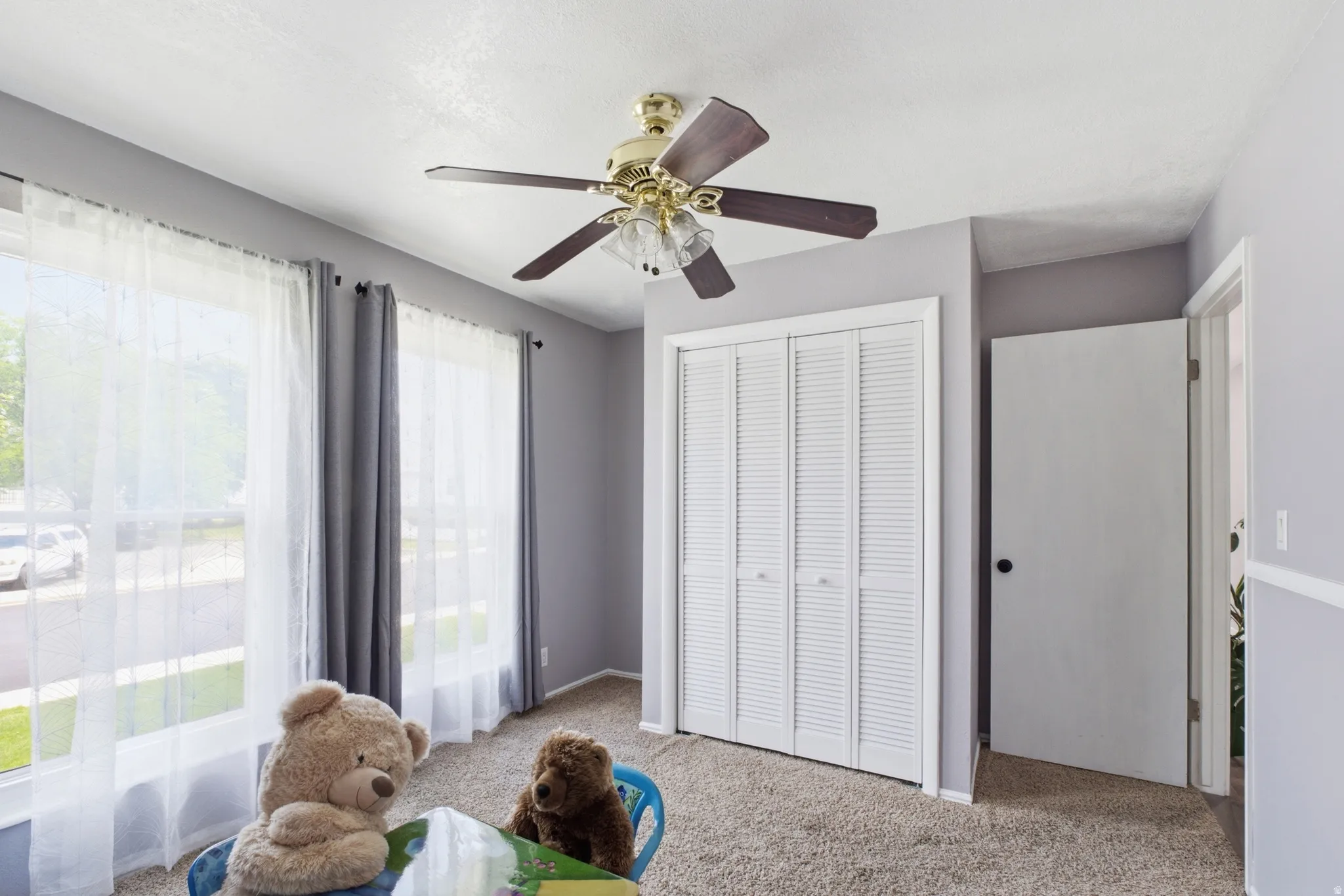 Light gray painted interior room featuring two large windows, a ceiling fan with light kit, bi-fold louvered closet doors, and carpet flooring