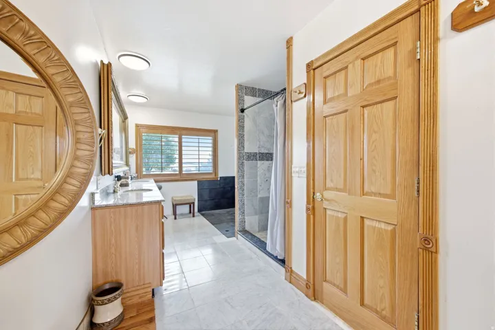 Main home - Expansive master Bathroom featuring a wood-panel door with decorative fluted trim, a double vanity with stone countertops and undermount sinks, a large mirror with an ornate carved wood frame, a window with wood plantation shutters, and a tile