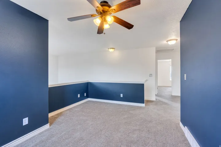 Carpeted loft area featuring a ceiling fan with integrated lighting, recessed wall paneling, white trim, and a hallway entry