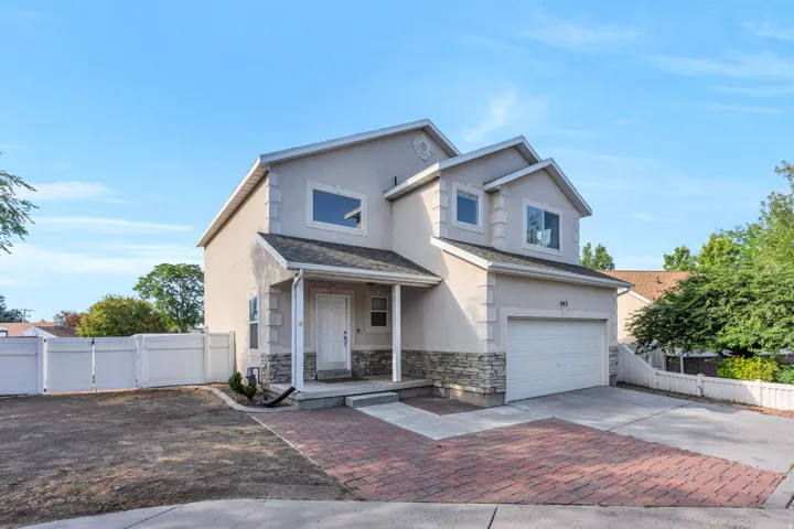 Two-story residential exterior featuring a stucco and stone facade, an attached garage, a covered front entry, a brick paver driveway, and white vinyl fencing