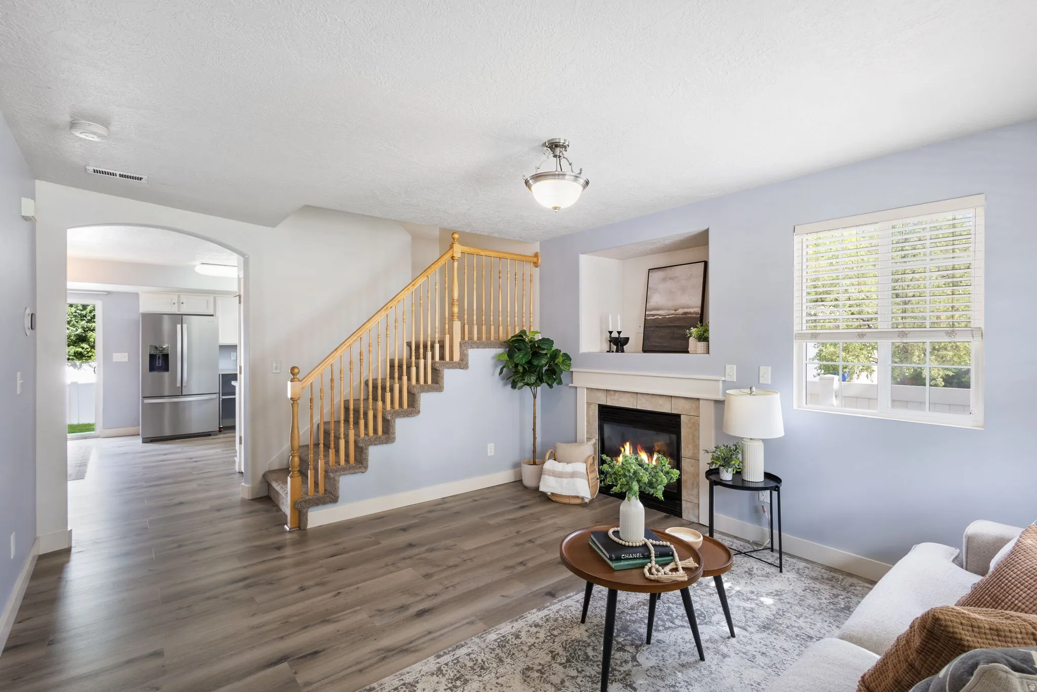 Living area featuring wood-finish flooring, a staircase with wood railing, fireplace with stone surround and mantle, and an arched doorway leading to a kitchen with stainless steel appliances