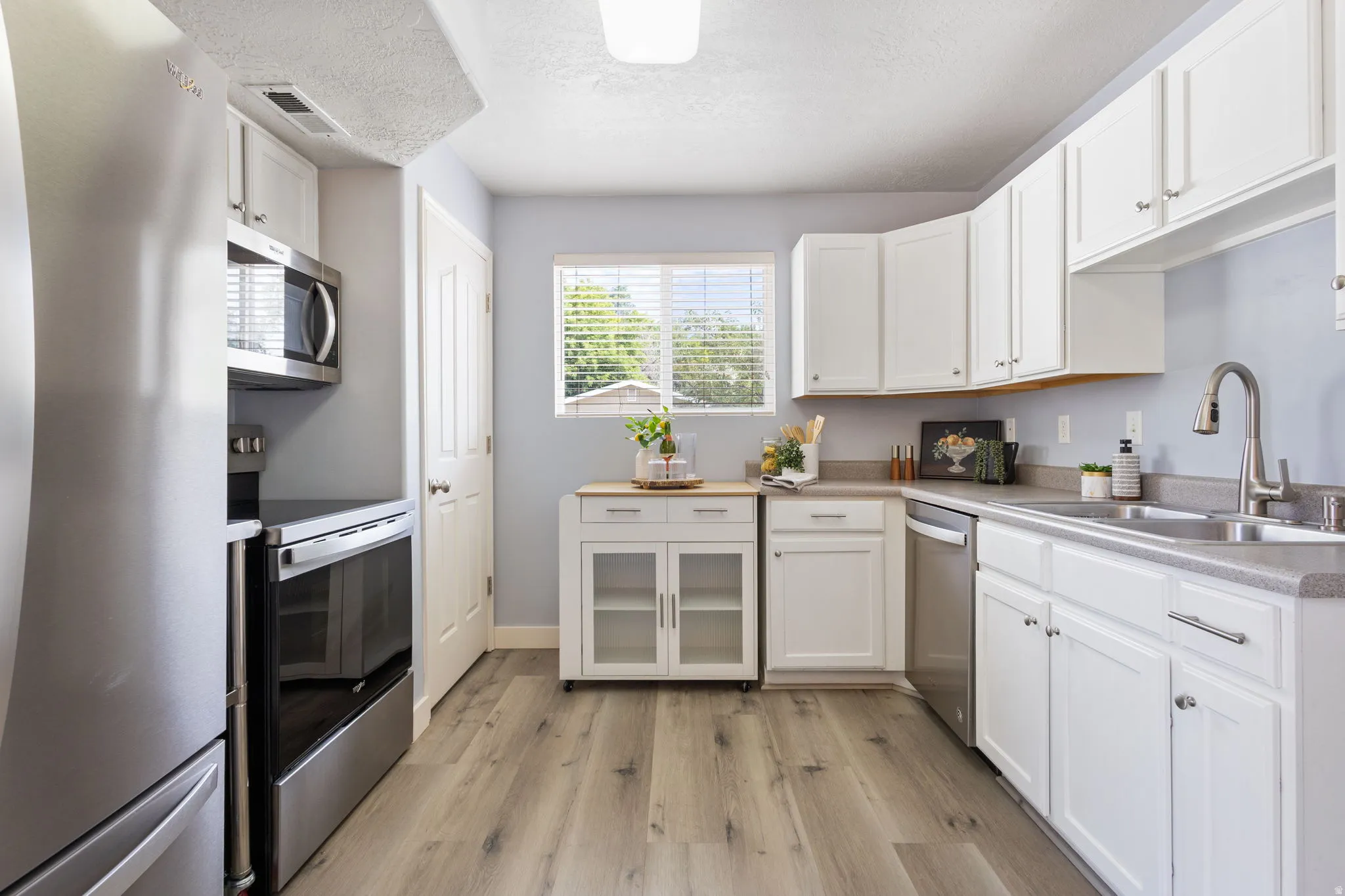 Modern kitchen featuring stainless steel appliances, white cabinetry, a double basin sink with a gooseneck faucet, light-toned countertops, and wood-finish flooring