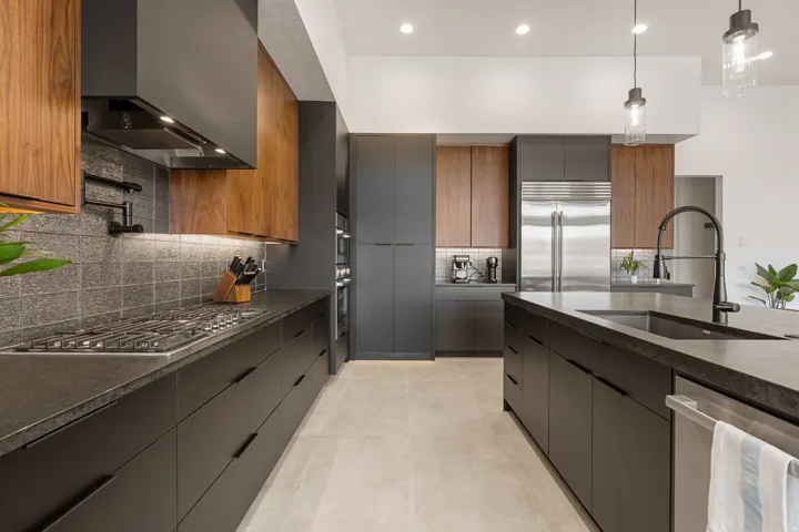 Contemporary kitchen featuring dark flat-panel cabinetry, wood-finish upper cabinets, stainless steel appliances, dark countertops, and a full-height tile backsplash