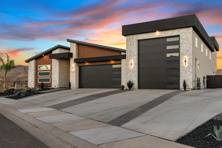 Contemporary architectural style featuring a multi-tone exterior, dark modern garage doors with horizontal windows, natural stone, wood-finish siding, and decorative exterior lighting