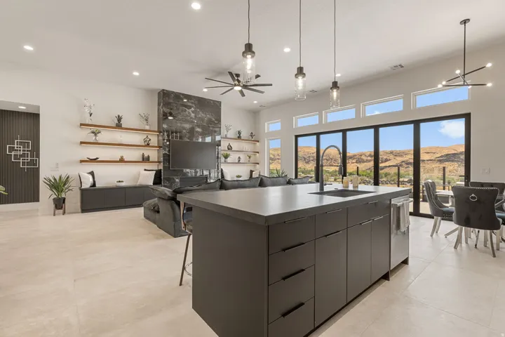 Expansive open-concept living area featuring a large center island with integrated sink, abundant natural light from floor-to-ceiling glass doors, a prominent dark-toned accent wall, recessed and pendant lighting, and light-toned tile flooring