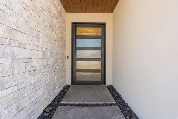 Contemporary entry featuring a dark-framed door with five frosted glass panels, stacked stone, a smooth stucco wall, wood-finish soffit, and a segmented concrete walkway accented with dark pebbles