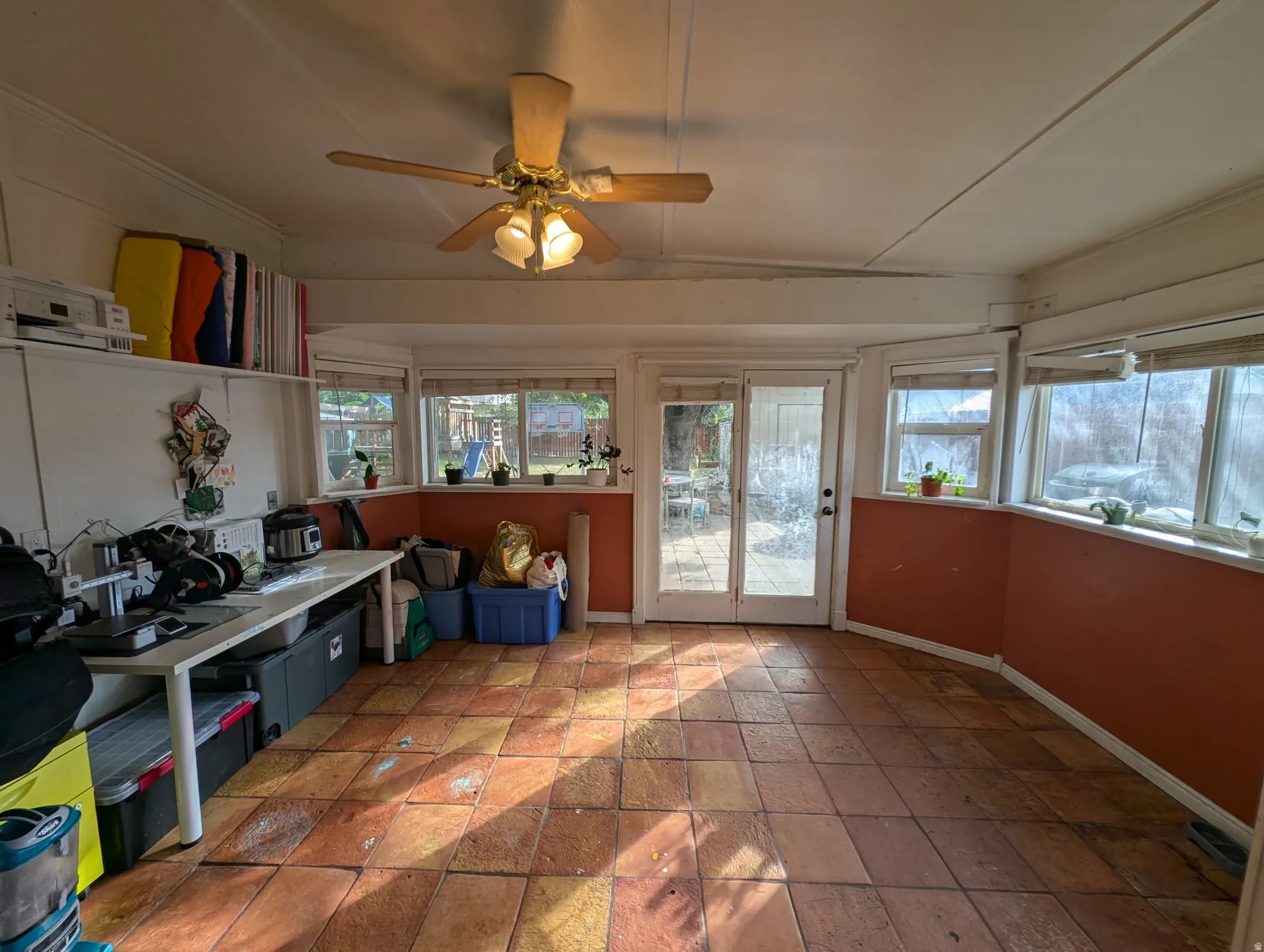 Terracotta-style tile flooring, multiple windows with natural light, glass-paned double doors, ceiling fan, and built-in shelving