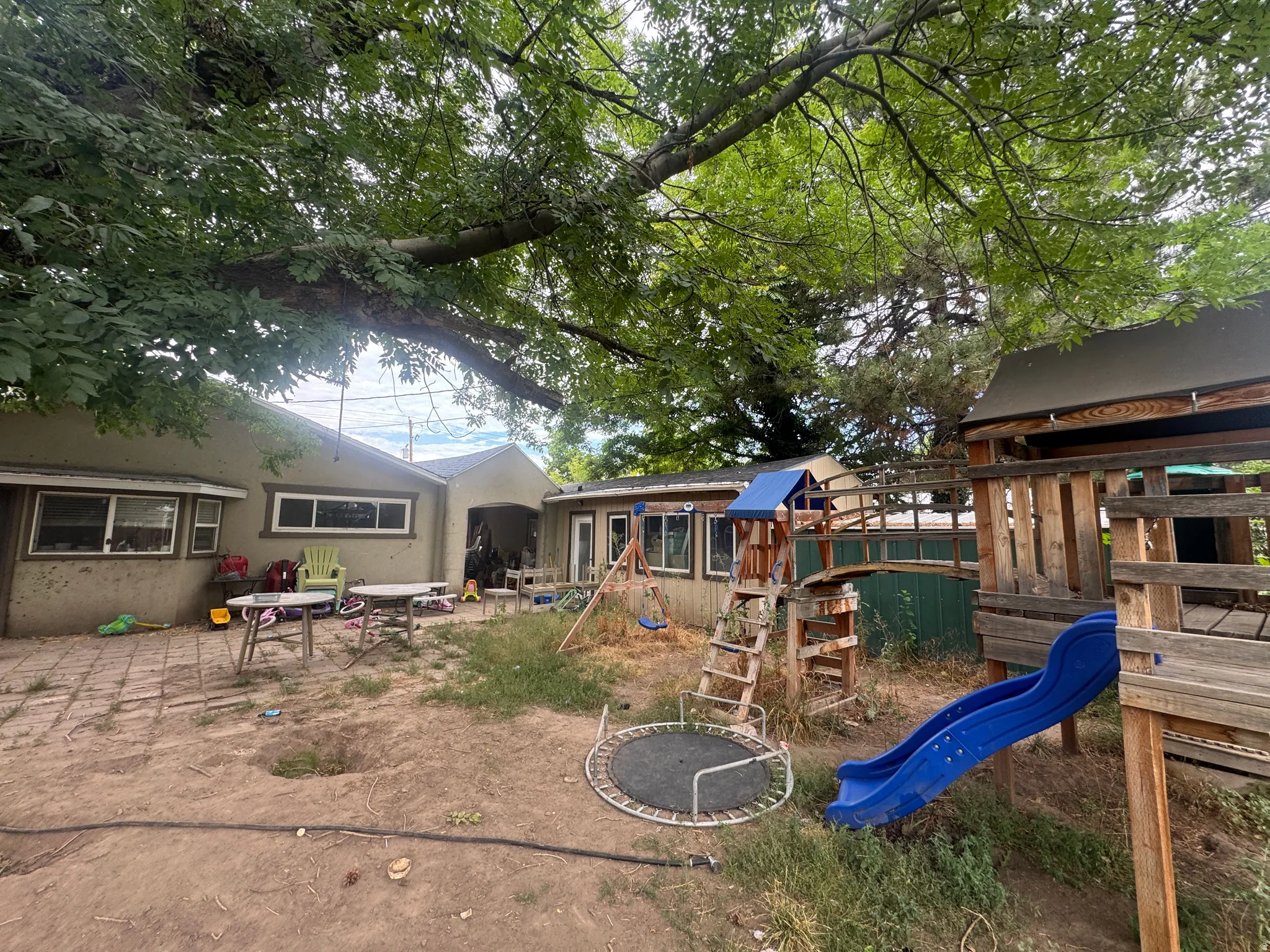 Expansive backyard featuring mature shade trees, a stone patio area, and a large wooden playset with a blue slide