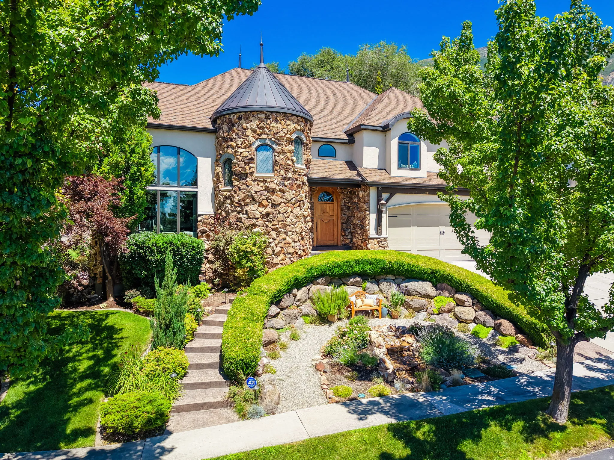 Stone-clad turret home with an arched wood entry door, two-car garage, and a tiered landscaped front yard featuring a stone staircase and rock garden