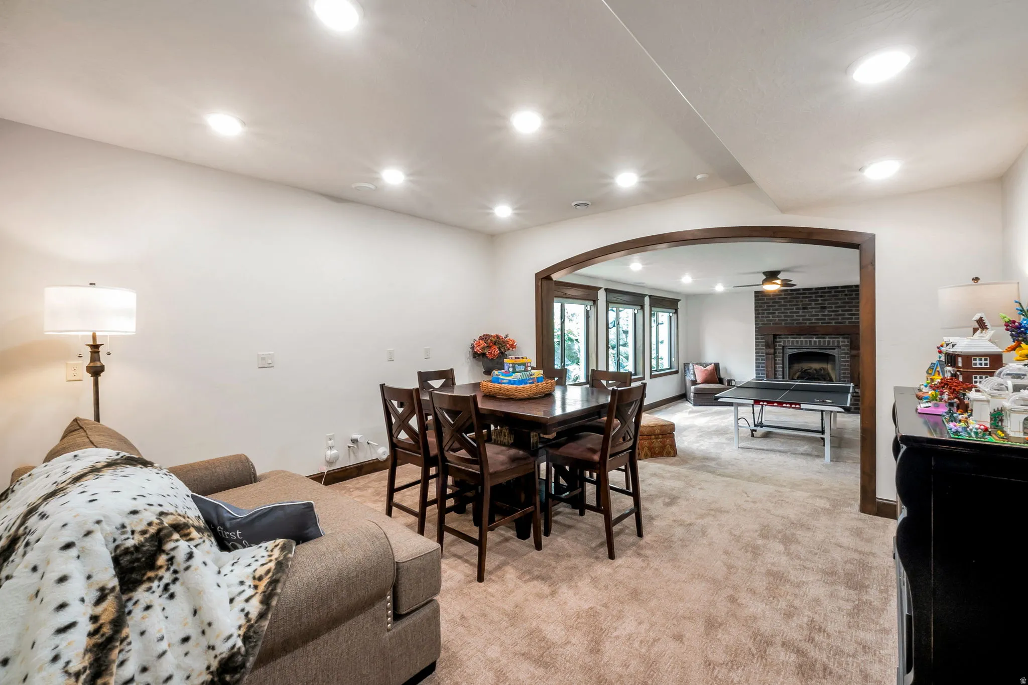 Open-concept recreation area featuring recessed lighting, carpet flooring, and an arched entryway to a separate sitting area with a brick fireplace and multiple windows