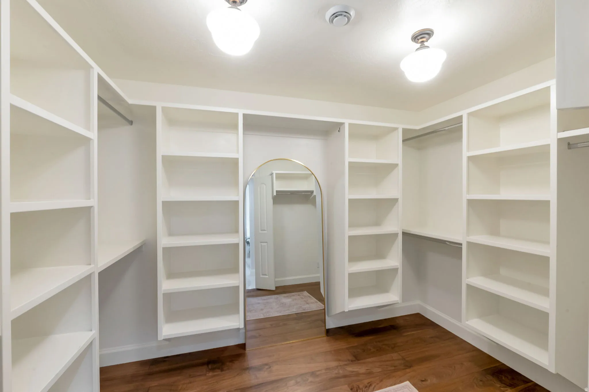 Built-in white shelving and hanging rods in a walk-in closet with dark wood-finish flooring, an arched doorway, and overhead dome lighting